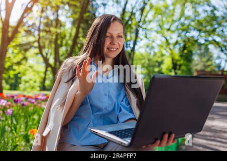 Außenporträt einer glücklichen Geschäftsfrau, die im Spring Park auf einem Laptop arbeitet. Eine Frau ruft per Videokonferenz an und spricht mit Cowokern im öffentlichen Garten Stockfoto