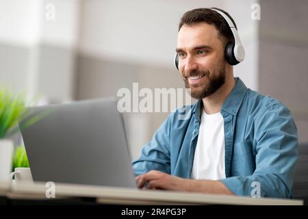Lächelnder männlicher Angestellter mit kabellosen Kopfhörern, der an einem Computer in einem modernen Büro arbeitet Stockfoto