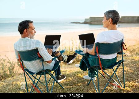Ältere Ehepartner, Die Online Auf Laptops In Der Nähe Des Ozeans Arbeiten, Rückansicht Stockfoto