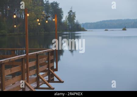 Vintage-Dekoration der Hochzeitszeremonie auf dem Holzpier. Terrasse dekoriert mit elektrischen Lampen als Girlande, Holzterrasse in der Nähe des Sees, Pier. Romantischer geldautomat Stockfoto