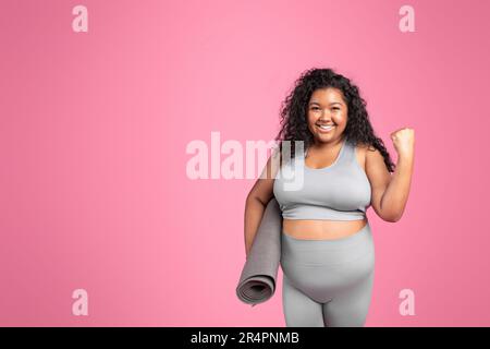 Aufgeregte schwarze, übergroße Dame in Sportbekleidung macht Siegesschild, feiert Erfolg, Haltematte, rosa Hintergrund, freier Platz Stockfoto