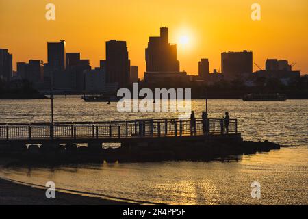 Blick von Odaiba, Tokio, über die Bucht bis zur Skyline von Tokio bei Sonnenuntergang mit Silhouetten von unbekannten Menschen im Vordergrund Stockfoto