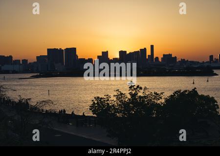 Blick von Odaiba, Tokio, über die Bucht bis zur Skyline von Tokio bei Sonnenuntergang mit Silhouetten von unbekannten Menschen im Vordergrund Stockfoto