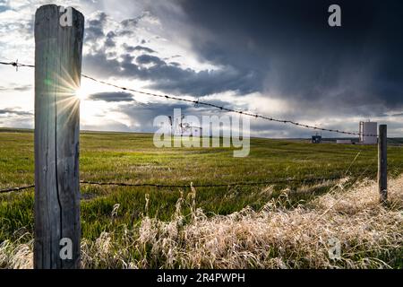 Landschaftskulisse in der Prärie mit Stacheldrahtzaun und Blick auf einen Ölpumpenbuckel unter stürmischem Himmel im Rocky View County Alberta, Kanada. Stockfoto