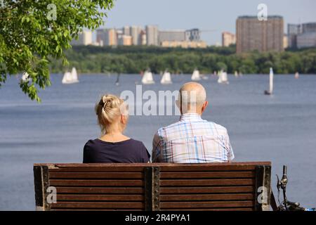 Alte Frau und Mann sitzen auf einer Holzbank am Fluss und im Hintergrund der Stadt, Rückblick. Ein paar Rentner am Strand, entspannen und das Leben im Sommer genießen Stockfoto