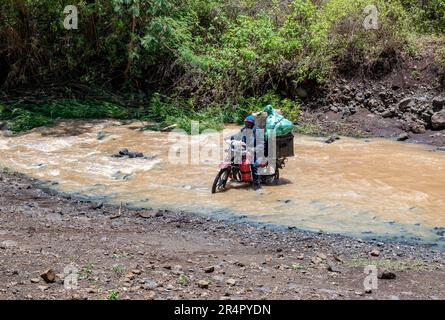 Ein Einheimischer auf einem schwer beladenen Motorrad, der versucht, einen Fluss zu überqueren. Kenia, Afrika. Stockfoto