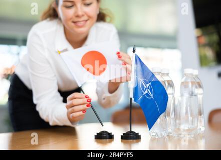 Mitarbeiter der Delegation bereitet Verhandlungstisch vor – richtet die Flagge der NATO (OTAN) und Japans ein Stockfoto