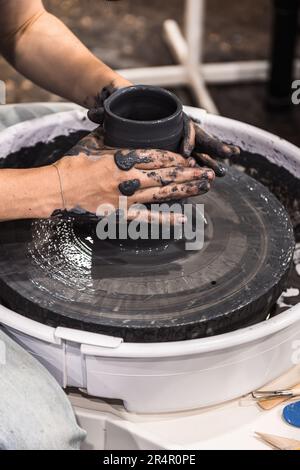 Eine Frau, die an einem Töpferrad arbeitet und mit Händen einen Becher mit dunkelschwarzem Lehm macht Stockfoto