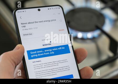 Die Hand eines Mannes mit einem Smartphone und einer E-Mail von British Gas über die neue Obergrenze für den Energiepreis im Juli 2023. Ein Gaskochfeld brennt im Hintergrund. UK Stockfoto