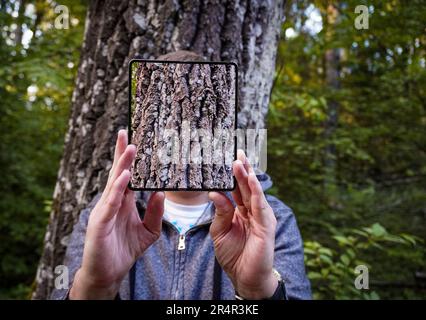 Mann, der ein digitales Tablet hält, während er gegen den Baumstamm steht Stockfoto