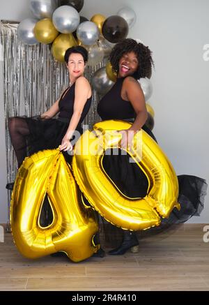 Zwei fröhliche Multirassenfrauen auf einer 40. Geburtstagsparty mit goldenen Ballons zum 40. Geburtstag. Stockfoto