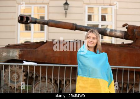 Junge Frau, in ukrainische Flagge eingewickelt, in der Nähe eines kaputten Panzers auf der Straße Stockfoto