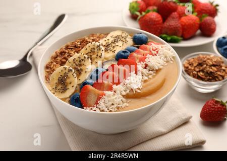 Köstliche Smoothie-Schüssel mit frischen Beeren, Banane, Kokosflocken und Müsli auf weißem Tisch, Nahaufnahme Stockfoto