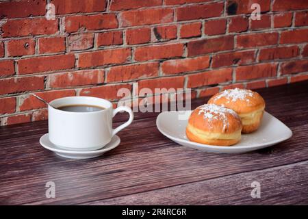 Eine Tasse Kaffee auf einer Untertasse mit einem Löffel und ein Teller mit frischen Donuts in Puderzucker auf einem Holztisch vor einer Ziegelwand. Nahaufnahme. Stockfoto