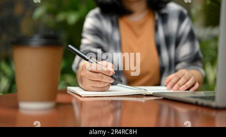 Zugeschnittenes Bild einer jungen asiatischen Studentin, die an ihren Aufgaben arbeitet und in einem Café im Freien Notizen auf ihrem Notizbuch schreibt. Stockfoto