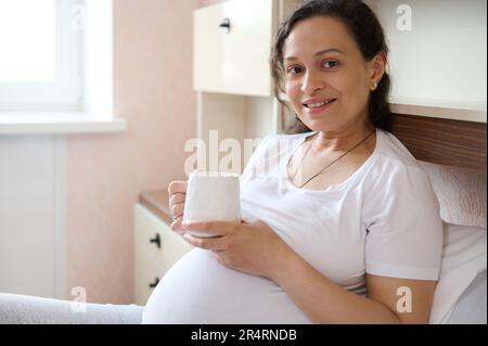 Schöne Erwachsene schwangere Frau, die lächelt und in die Kamera schaut, sich auf dem Bett entspannt und eine große Tasse gesundes Getränk hält. Stockfoto