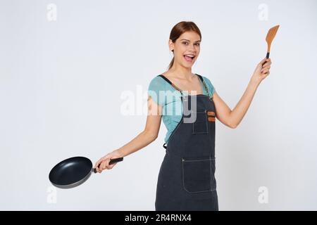 Junge Frau trägt Küchenschürze und kocht und hält Pfanne und Spachtel isoliert auf weißem Hintergrund. Stockfoto