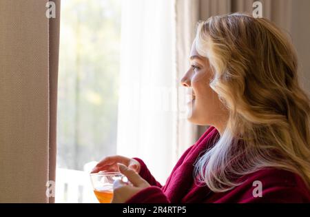 Lächelnde, übergroße, bedachtsame junge Frau mit Teetasse, die zu Hause durch das Fenster blickt Stockfoto