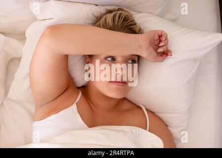 Blick aus einem hohen Winkel auf eine übergroße, bedachtsame, weiße junge Frau, die auf einem gemütlichen Bett liegt und wegschaut Stockfoto