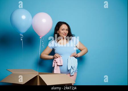Glückliche schwangere Frau, die Neugeborene-Kleidung auf dem Bauch hält und mit pinkfarbenen und blauen Ballons auf blauem Hintergrund posiert Stockfoto