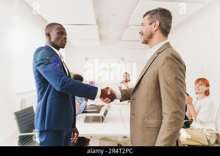 Multicultural Office Handshake - ein Afrikaner und ein Weißer schütteln in einem modernen Büro die Hand, während Kollegen im Hintergrund applaudieren. Stockfoto