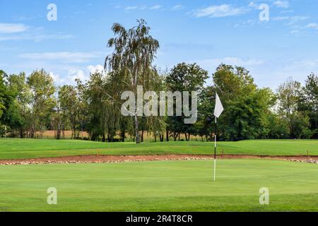 Malerischer Blick auf grünes Gras auf dem Golfplatz mit weißer Flagge vor üppigen Bäumen und bewölktem blauem Himmel Stockfoto