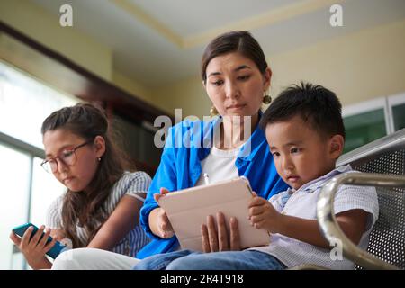 Mutter und Kinder schauen sich unterhaltsame Videos auf Geräten an, während sie im Krankenhaus in der Schlange stehen Stockfoto