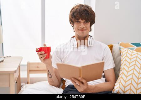 Junger blonder Mann liest Buch und trinkt Kaffee im Schlafzimmer Stockfoto