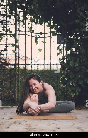 Fröhliche Yoga-Lehrerin mit einem Bein, nach vorne gefaltet, nach vorne gebeugt, auf die Kamera gelächelt, langes brünette Haar, grünes Outfit, Korkmatte, draußen, Pflanzen an Stockfoto