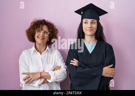 Hispanische Mutter und Tochter, die eine Abschlussmütze und einen Morgenmantel tragen, lächelnd zur Seite schauen und wegschauen und nachdenken. Stockfoto