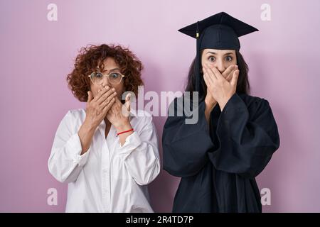 Hispanische Mutter und Tochter tragen Abschlussmütze und zeremoniellen Morgenmantel, der den Mund mit Händen verdeckt. Geheimes Konzept. Stockfoto