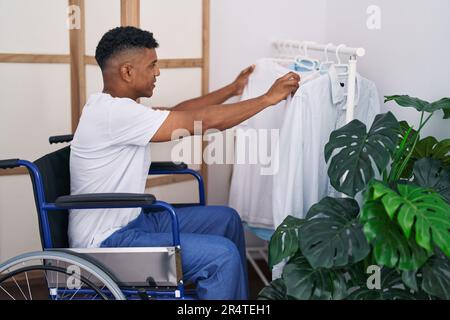 Ein junger lateiner, der ein Hemd aussucht, sitzt im Rollstuhl im Schlafzimmer Stockfoto