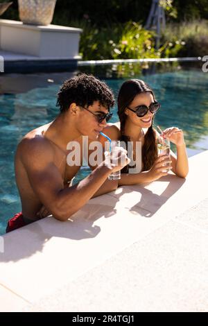 Ein birassisches junges Paar mit Sonnenbrille, das an sonnigen Tagen im Swimmingpool entspannt einen Drink zu sich nimmt Stockfoto