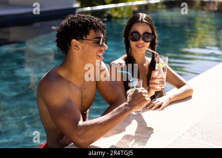 Ein birassisches junges Paar mit Sonnenbrille, das an sonnigen Tagen im Pool einen Drink zu sich nimmt Stockfoto