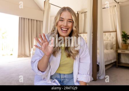Porträt einer glücklichen jungen weißen Frau, die im Schlafzimmer mit der Hand winkt Stockfoto