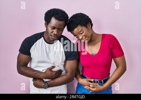 Junges afroamerikanisches Paar, das über rosa Hintergrund steht, mit Hand auf Bauch, weil Verdauungsstörungen, schmerzhafte Krankheit sich unwohl fühlt. Ache-Konzept. Stockfoto