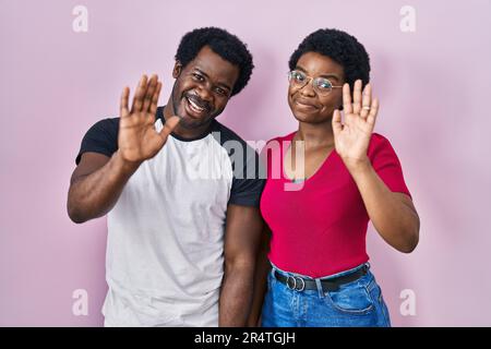 Ein junges afroamerikanisches Paar, das über rosa Hintergrund steht und darauf verzichtet, Hallo zu sagen, glücklich und lächelnd, freundliche Willkommensgeste Stockfoto