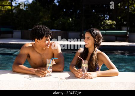 Ein birassisches junges Paar mit Getränken, das sich gegenseitig anschaut, während es am sonnigen Tag im Swimmingpool genoss Stockfoto