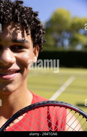 Nahaufnahme eines birassischen jungen Mannes mit Tennisschläger, der am sonnigen Tag auf dem Tennisplatz lächelt Stockfoto