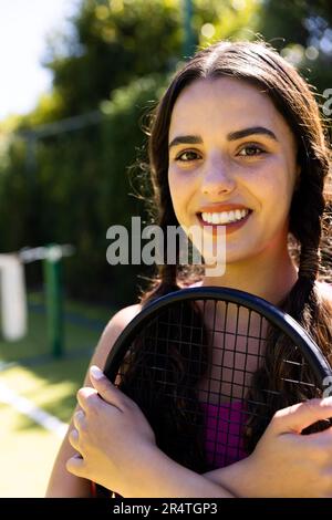 Nahaufnahme einer lächelnden, wunderschönen, birassischen jungen Frau mit Tennisschläger auf dem Tennisplatz am sonnigen Tag Stockfoto
