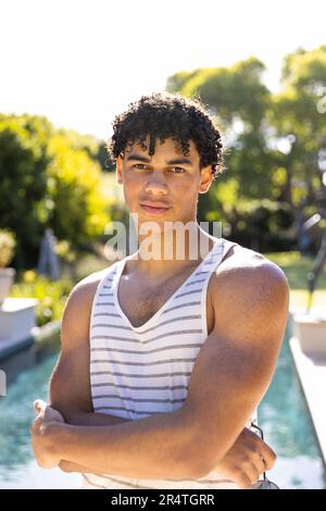 Porträt eines birassischen jungen Mannes, der eine Sonnenbrille hält und am Pool vor klarem Himmel steht Stockfoto