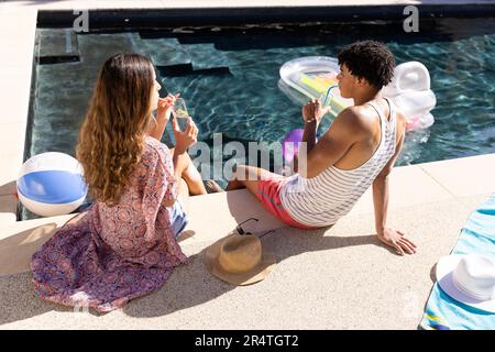Blick aus einem hohen Winkel auf ein birassisches junges Paar, das Limonade trinkt, während man am Pool sitzt Stockfoto
