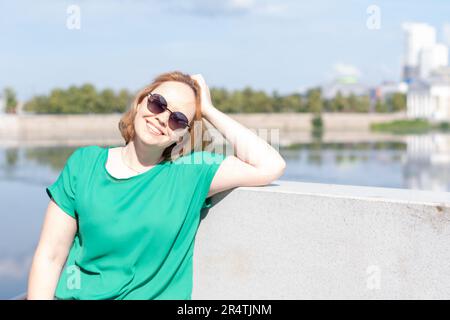 Porträt eines Mädchens mit Sonnenbrille, eine grüne Bluse, die am Flussufer steht und ihr Haar mit der Hand hält, Kopierraum. Stockfoto