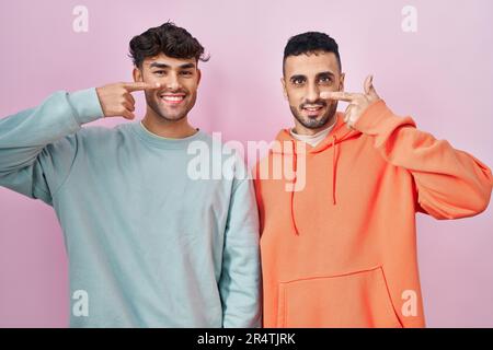 Ein junges, lateinamerikanisches, schwules Paar, das auf pinkfarbenen Hintergrund steht und mit der Hand auf Gesicht und Nase zeigt, lächelt fröhlich. Schönheitskonzept Stockfoto