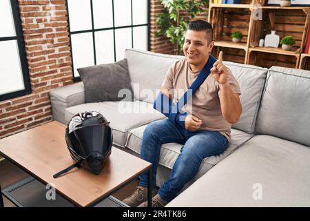 Hispanischer junger Mann, der den Arm an der Schlinge trägt, wegen eines Motorradunfalls überrascht von einer Idee oder einer Frage, mit dem Finger auf ein glückliches Gesicht zu zeigen, Nummer eins Stockfoto