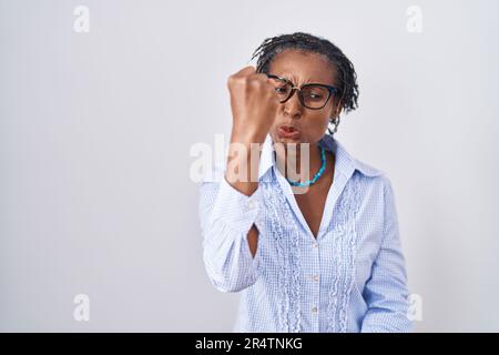 Afrikanische Frau mit Dreadlocks, die auf weißem Hintergrund steht und eine Brille trägt, wütend und verrückt, die Faust erhebt, frustriert und wütend, während sie mit Ang schreit Stockfoto