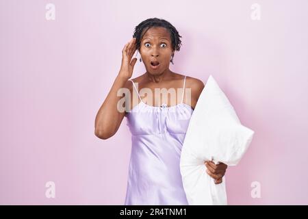 Afrikanische Frau mit Dreadlocks, die Pyjama trägt und Kissen umarmt, verrückt und verängstigt mit Händen am Kopf, ängstlich und überrascht von Schock mit offenem Mund Stockfoto