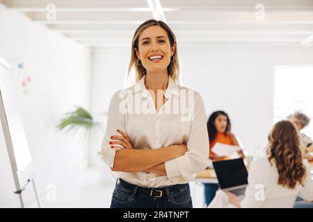 Glückliche junge Geschäftsfrau, die vor der Kamera lächelt, während sie in einem Konferenzraum steht. Fröhliche junge Geschäftsfrau, die an einem Treffen mit ihren Kollegen teilnimmt Stockfoto