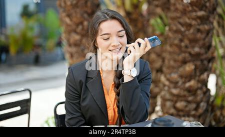 Eine junge, wunderschöne hispanische Geschäftsfrau, die eine Sprachnachricht per Smartphone sendet und auf der Terrasse des Cafés lächelt Stockfoto