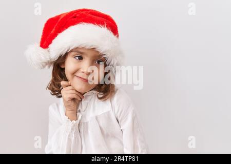 Ein kleines lateinamerikanisches Mädchen, das einen weihnachtsmütze trägt und über Zweifel mit dem Finger am Kinn nachdenkt und sich wundert Stockfoto